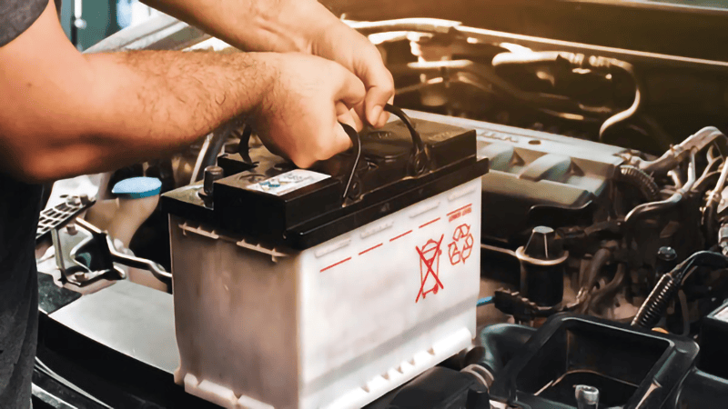Close-up shot of hands installing or handling a large car battery within an engine bay.