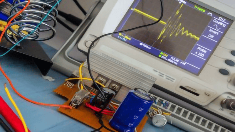 Close-up of an electronic experimental setup: An oscilloscope (displaying a yellow waveform on its screen) connected to a breadboard with components like a blue capacitor, colored wires, and a coiled inductor placed nearby.