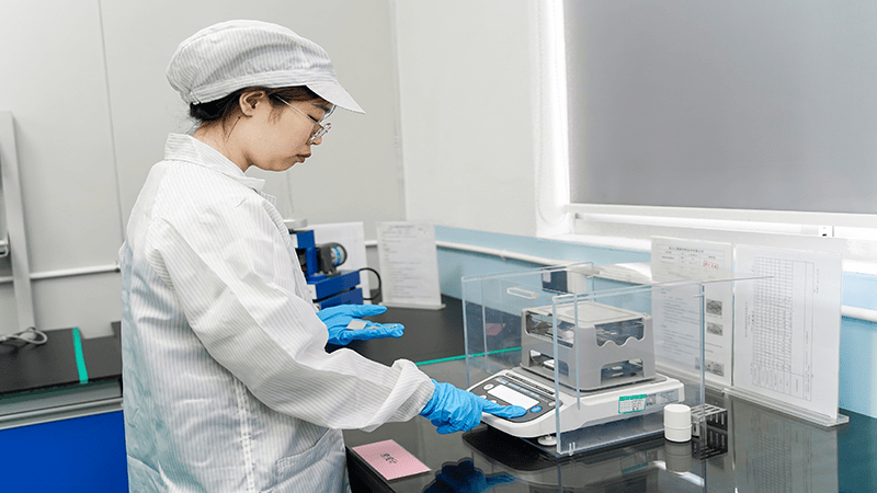 Female lab tech in cleanroom gear operates a precision analytical balance (in a transparent case) on a lab bench, in a sterile lab setting.
