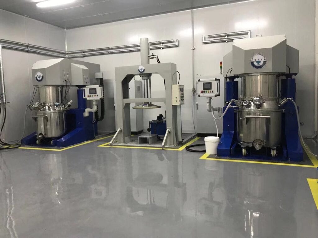 Cleanroom facility with multiple industrial planetary mixing machines (stainless steel construction) — featuring control panels, blue/gray components, a smooth glossy gray floor marked with yellow safety lines, and wall-mounted pipes. These machines are typically used for blending materials like coatings or battery slurries.