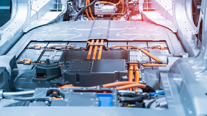 Interior view of an electric vehicle’s battery pack installed in the car chassis, with bright orange high-voltage cables connecting the battery modules and adjacent vehicle components.