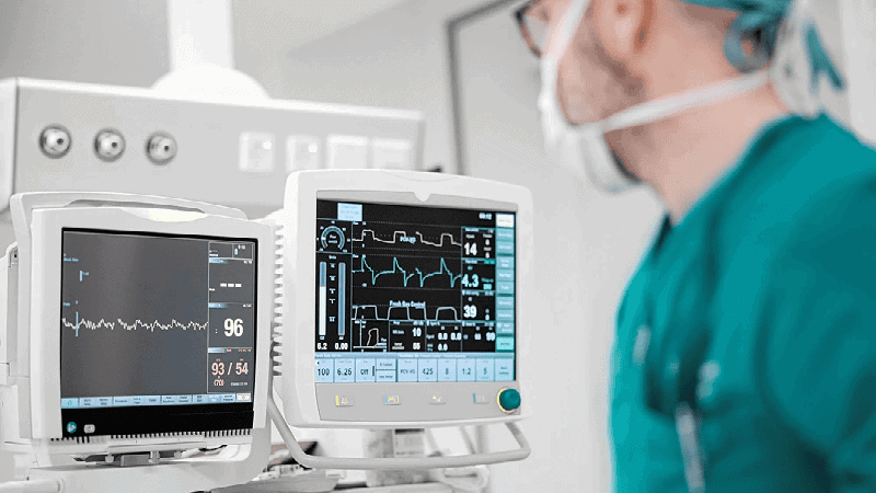 Monitores de signos vitales médicos que muestran datos del paciente en un quirófano, con un cirujano en uniforme médico cerca.
