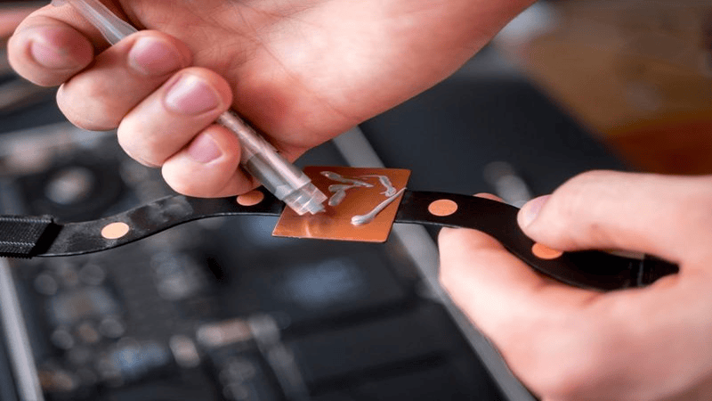 Solder paste application on flexible PCB: Hands dispensing solder paste onto a copper pad on a black flexible circuit board, a key step in electronics assembly and printed circuit board (PCB) manufacturing
