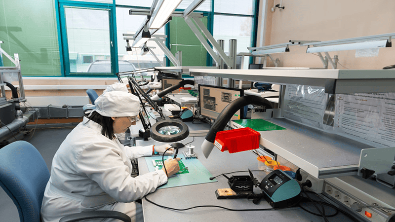Technician in a cleanroom suit assembling or repairing an electronic circuit board at a workbench