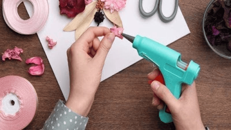 Person using a hot glue gun to attach pink decorative flowers for a DIY craft project.